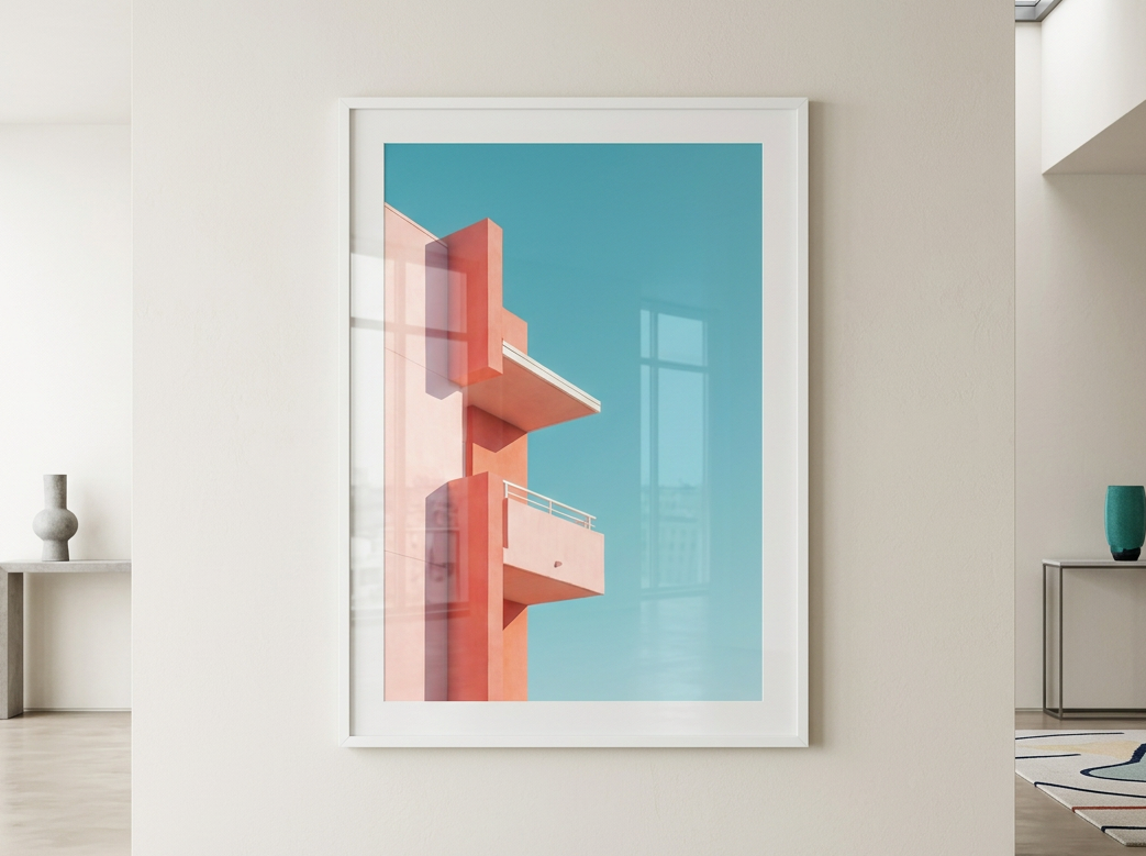 A framed art print featuring a minimalist geometric building design in coral pink and peach tones against a bright turquoise sky, displayed on a clean white wall in a modern minimalist room.