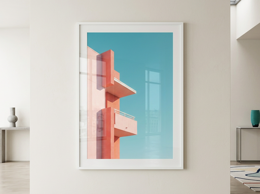 A framed art print featuring a minimalist geometric building design in coral pink and peach tones against a bright turquoise sky, displayed on a clean white wall in a modern minimalist room.