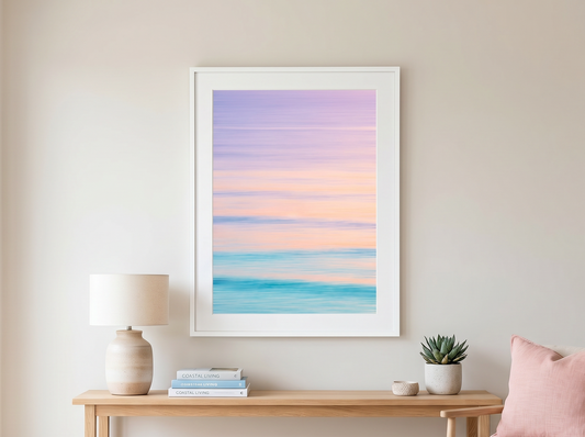A framed pastel ocean sunset print hangs on a beige wall above a light wood console table in a bright, modern room with a lamp, books, a small plant, and a pink cushion.