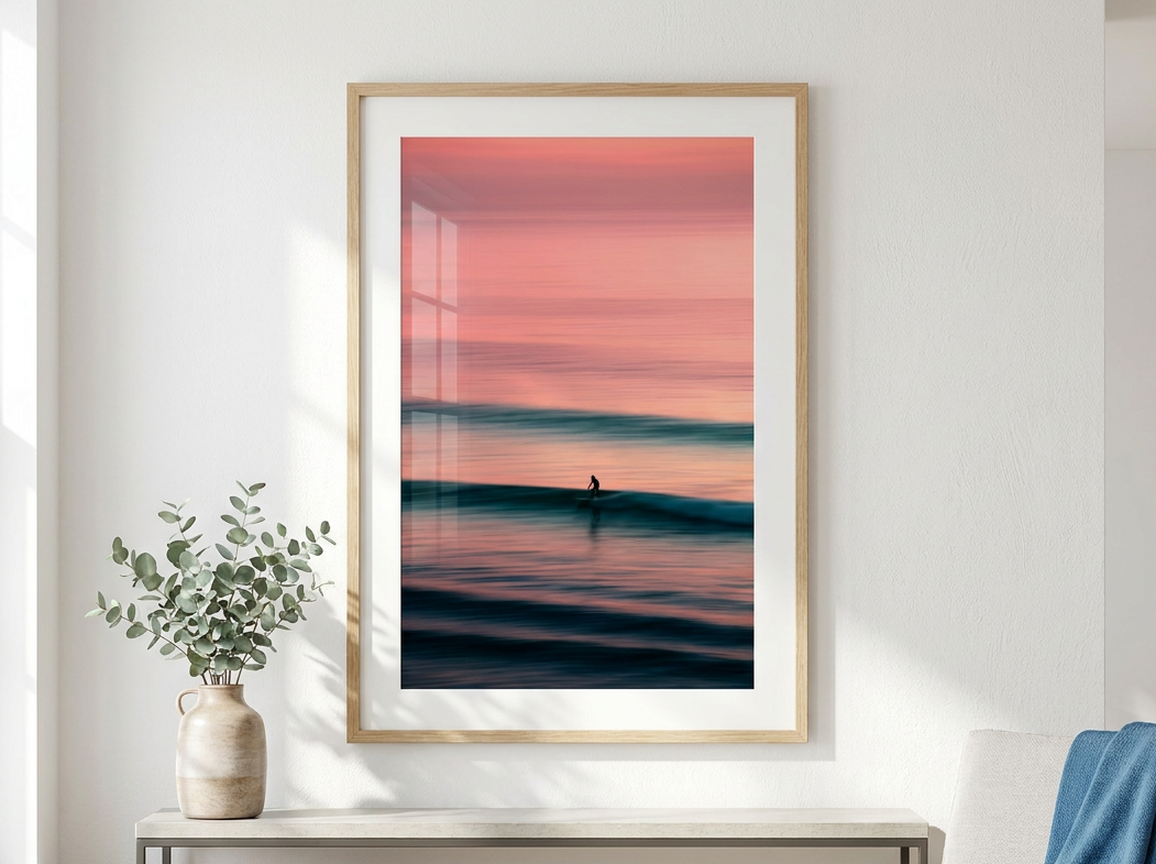 A framed vertical art print depicting a serene sunset over calm ocean waves, styled in a bright, modern living room setting above a console table.